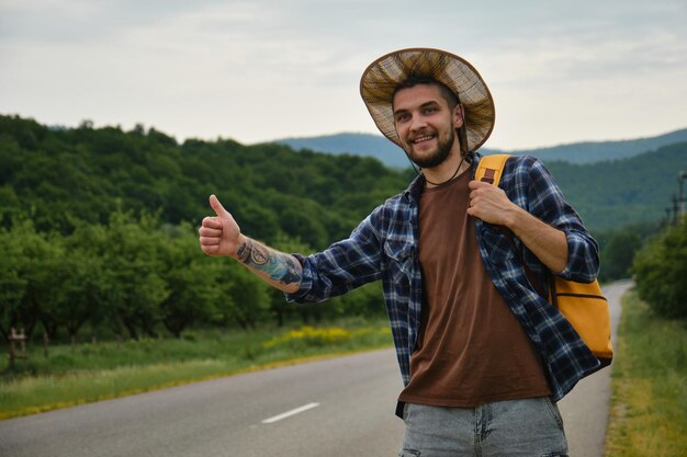 Hipster with hat and yellow backpack tries to stop car with thumbs up Concept of traveling alone