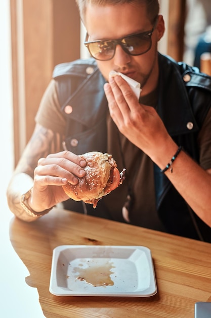 Hipster with a stylish haircut and beard sits at a table, decided to dine at a roadside cafe, eating a hamburger.