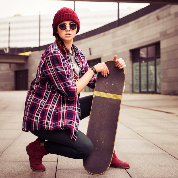 Foto donna hipster in abito casual seduto sul pavimento in città con lo skateboard nelle sue mani