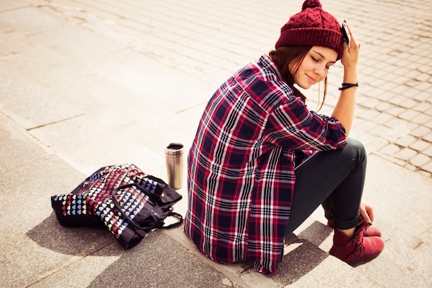 hipster woman in casual outfit sitting on steps in city with a phone in her hand