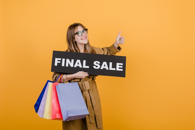 Hipster woman in glasses and coat with final sale sign and colorful shopping bags isolated over yellow