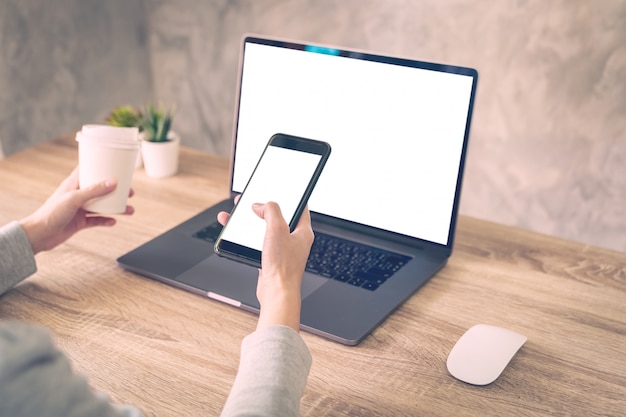 Hipster woman holding phone and using laptop on wooden table in coffee shop.