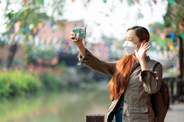 Photo hipster woman traveler with mask and backpack holding telephone making selfies speaking skype waving palm saying hi