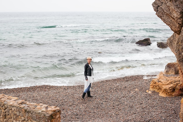 Хипстерская женщина в платье и куртке гуляет у моря осенью