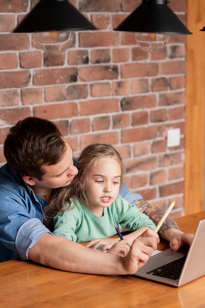 Hipster young father with tattoo on arm sitting at table with daughter and using laptop while helping her to prepare for school test