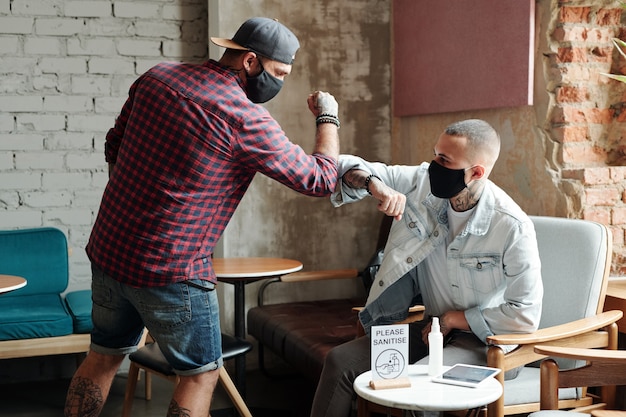 Hipster young friends in masks touching elbows instead of handshake during coronavirus while meeting in lobby