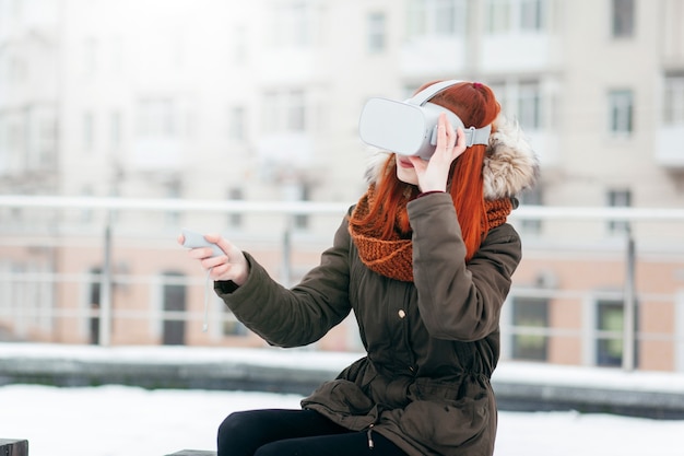 Hipster young girl plays virtual reality mobile game app on smartphone attached to modern VR glasses outdoor