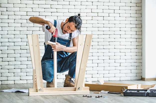 Hipster young man in apron working as handyman, assembling wood table with equipments, concept for home diy and self service.