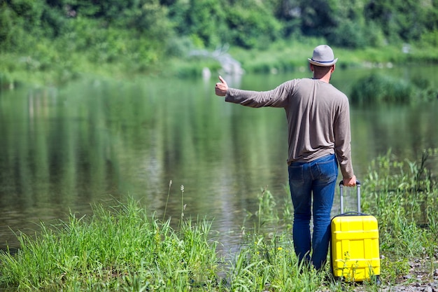 Hipster young man hitchhiking along a mountain river with a yellow suitcase