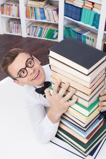 His favorite books. top view of excited young nerd man in shirt
and bow tie sitting at the table in library and hugging a book
stack