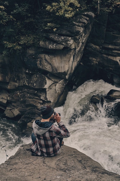 His favorite hobby. Top view of young modern man photographing while sitting on the rock with the river below