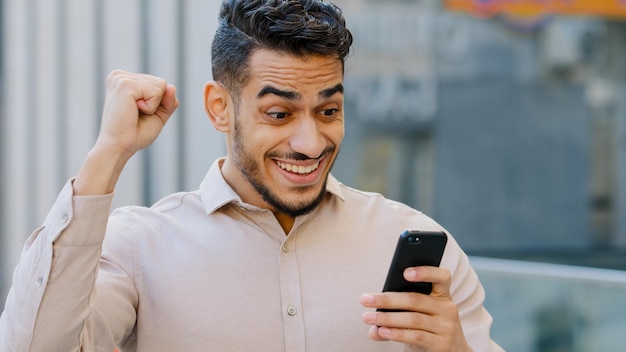 Uomo d'affari stupito ispanico che riceve messaggio sms offre opportunità uomo felice arabo che legge buone notizie in smartphone eccitato vincitore maschio felicissimo all'aperto che celebra il successo vittoria del telefono cellulare