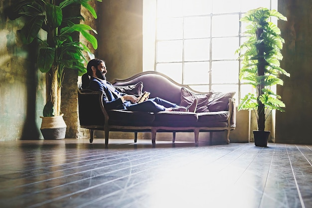 Испаноязычный бородатый бизнесмен расслабляется с помощью планшета, лежащего на диване в задней части офиса, деловых людей и технологической концепции