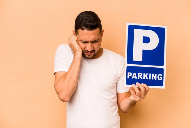 Hispanic caucasian man holding parking placard isolated on beige background covering ears with hands