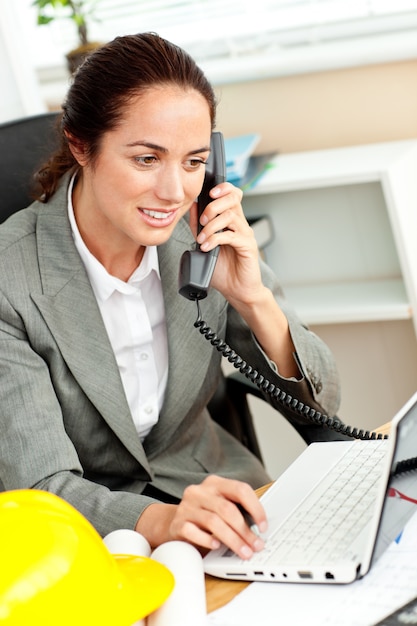 Architetto femminile ispano che per mezzo del suo computer portatile mentre parlando sul telefono