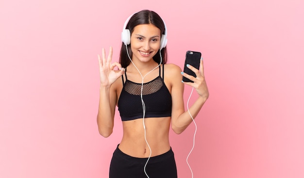 Donna ispanica fitness sentirsi felice, mostrando approvazione con un gesto ok con le cuffie e un telefono