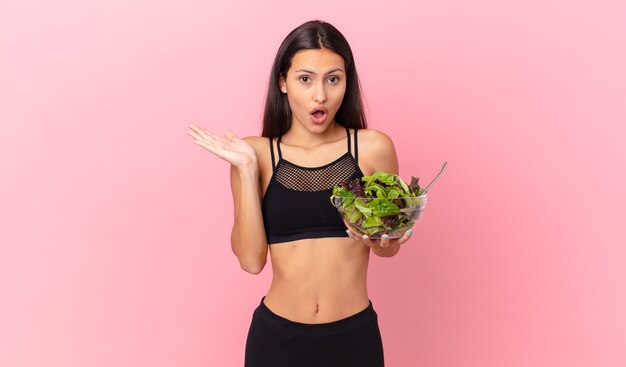 Hispanic fitness woman looking surprised and shocked, with jaw dropped holding an object and holding a salad