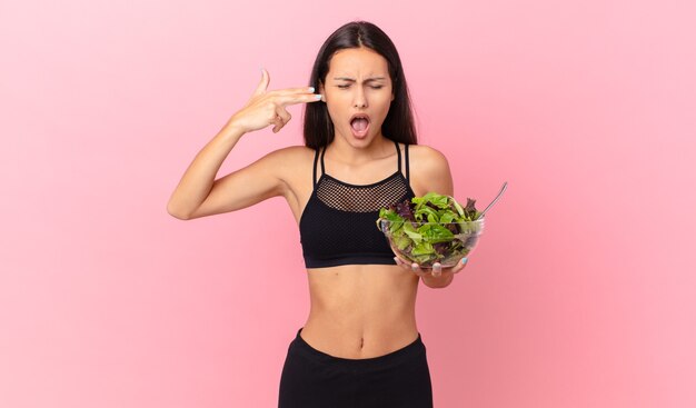 hispanic fitness woman looking unhappy and stressed, suicide gesture making gun sign and holding a salad