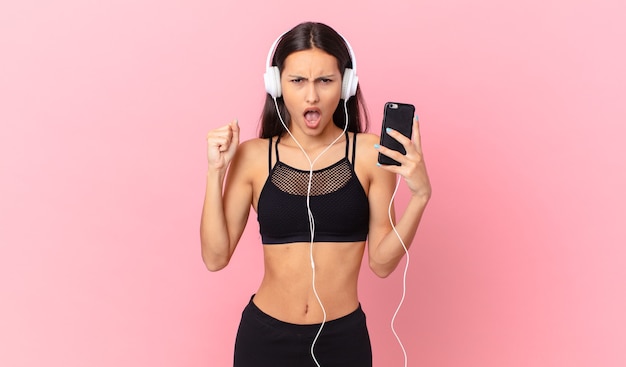 Hispanic fitness woman shouting aggressively with an angry expression with headphones and a phone