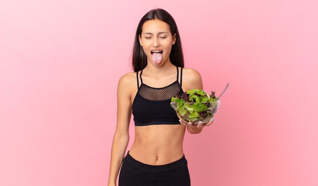 Hispanic fitness woman with cheerful and rebellious attitude, joking and sticking tongue out and holding a salad