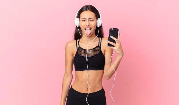 Hispanic fitness woman with cheerful and rebellious attitude, joking and sticking tongue out with headphones and a phone