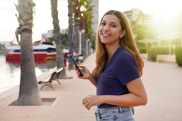 Hispanic girl smiling at camera holding smartphone on promenade