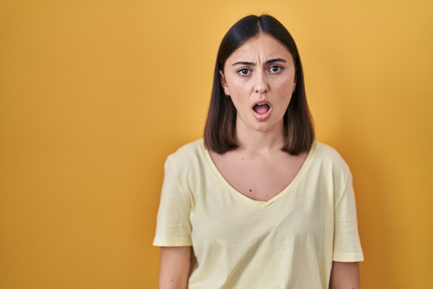 Hispanic girl wearing casual t shirt over yellow background in shock face looking skeptical and sarcastic surprised with open mouth