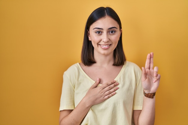 Hispanic girl wearing casual t shirt over yellow background smiling swearing with hand on chest and fingers up making a loyalty promise oath