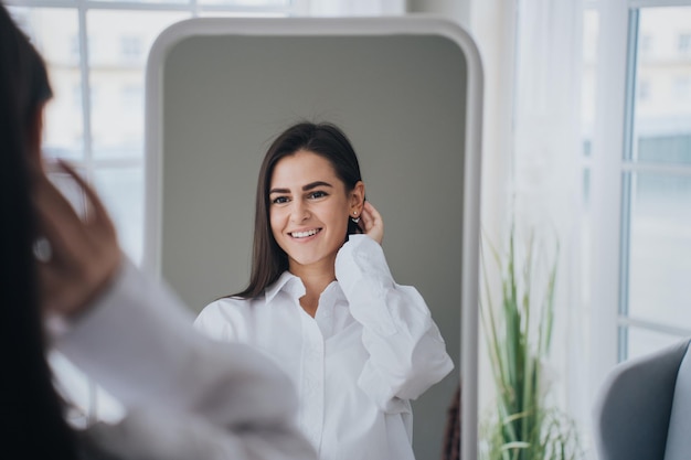 Hispanic gorgeous young woman in white shirt at home looking at mirror toothy smiling satisfied