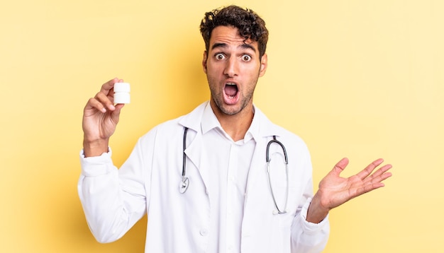 Photo hispanic handsome man amazed, shocked and astonished with an unbelievable surprise. physician bottle pills concept