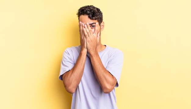 Hispanic handsome man covering face with hands, peeking between fingers with surprised expression