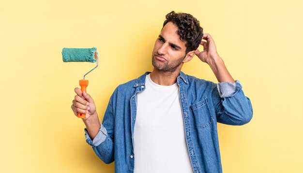 Hispanic handsome man feeling puzzled and confused, scratching head. roller paint concept