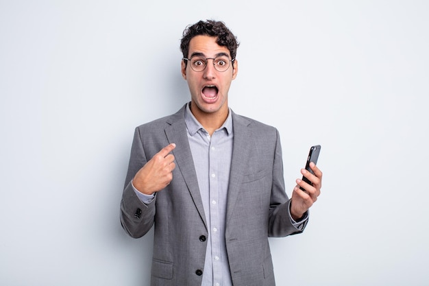 Hispanic handsome man looking shocked and surprised with mouth wide open, pointing to self. business and phone concept