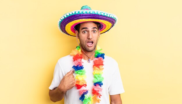 Hispanic handsome man looking shocked and surprised with mouth wide open, pointing to self. mexican party concept