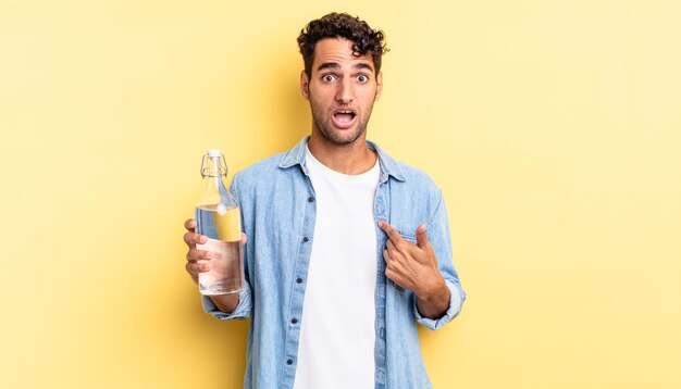 Hispanic handsome man looking shocked and surprised with mouth wide open, pointing to self. water bottle concept