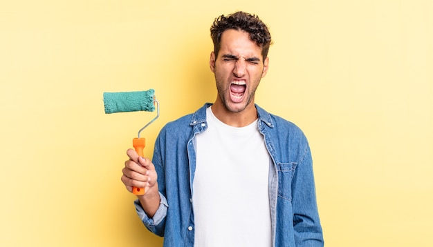 Hispanic handsome man shouting aggressively, looking very angry. roller paint concept