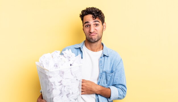 Hispanic handsome man shrugging, feeling confused and uncertain. paper balls trash concept