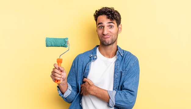 Hispanic handsome man shrugging, feeling confused and uncertain. roller paint concept