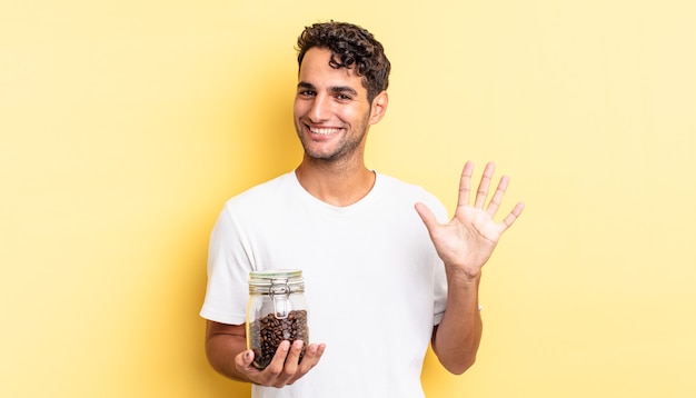 Hispanic handsome man smiling and looking friendly, showing number five. coffee beans bottle