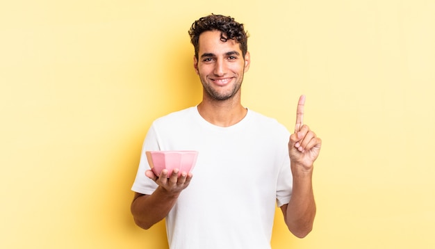 Hispanic handsome man smiling and looking friendly, showing number one. empty bowl concept