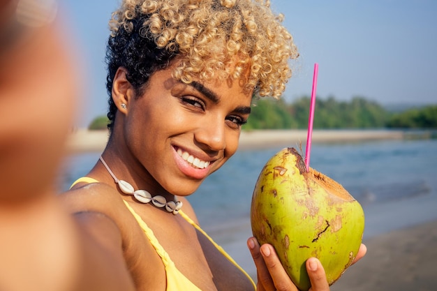 Hispanic latin beautiful female drinking coco nut water seashells on the neck and taking selfie on camera phone