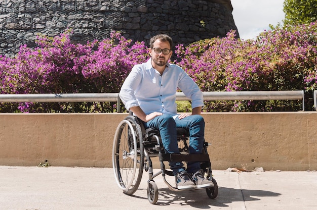 Hispanic Latino white adult male in a wheelchair outside in a park looking at the camera, with a nice garden behind him, on a sunny day.