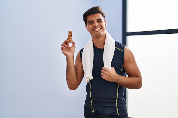 Hispanic man eating protein bar as healthy energy snack looking positive and happy standing and smiling with a confident smile showing teeth