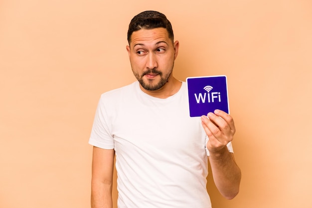 Hispanic man holding wifi placard isolated on beige background confused feels doubtful and unsure