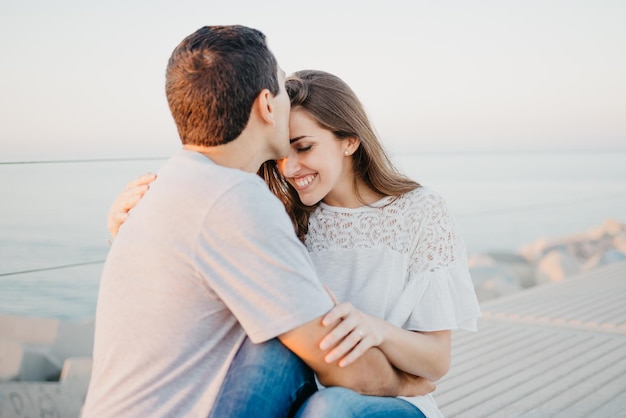 Hispanic man and a smiling brunette girl are sitting on a breakwater in Spain