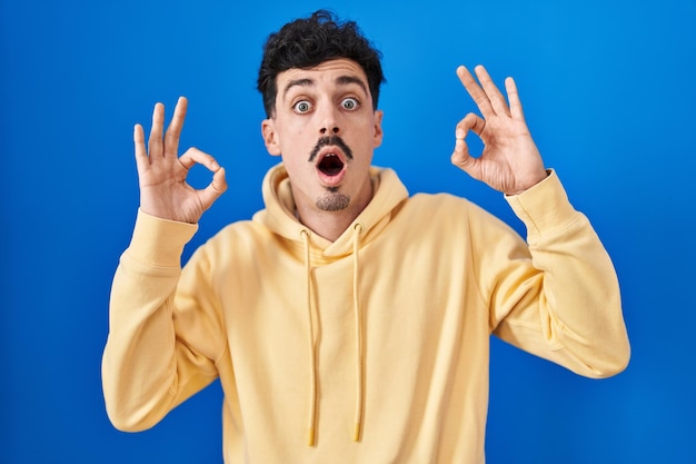 Hispanic man standing over blue background looking surprised and shocked doing ok approval symbol with fingers. crazy expression