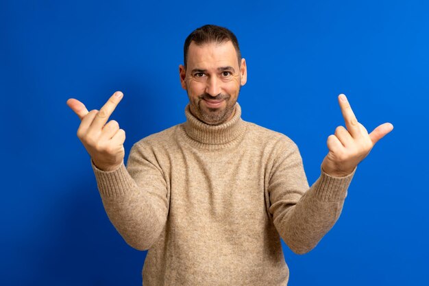 Hispanic man standing over blue background showing middle finger making fuck you bad expression provocation and rude attitude person