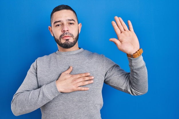 Hispanic man standing over blue background swearing with hand on chest and open palm, making a loyalty promise oath