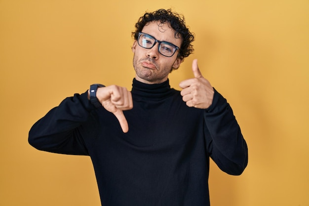 Hispanic man standing over yellow background doing thumbs up and down, disagreement and agreement expression. crazy conflict