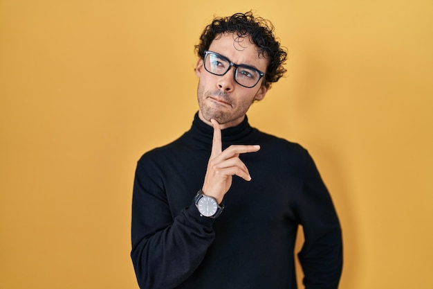 Hispanic man standing over yellow background thinking concentrated about doubt with finger on chin and looking up wondering